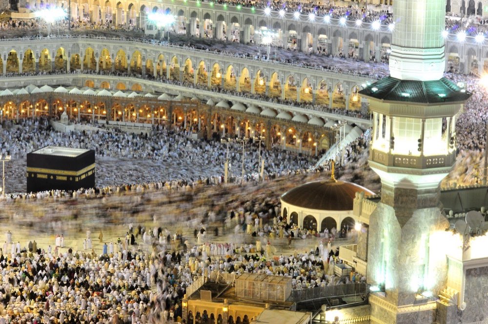 The farewell tawaf at night, photographed in long exposure: the Kaaba sharp at the centre and the circling crowd around it drawn out into a continuous band of movement.